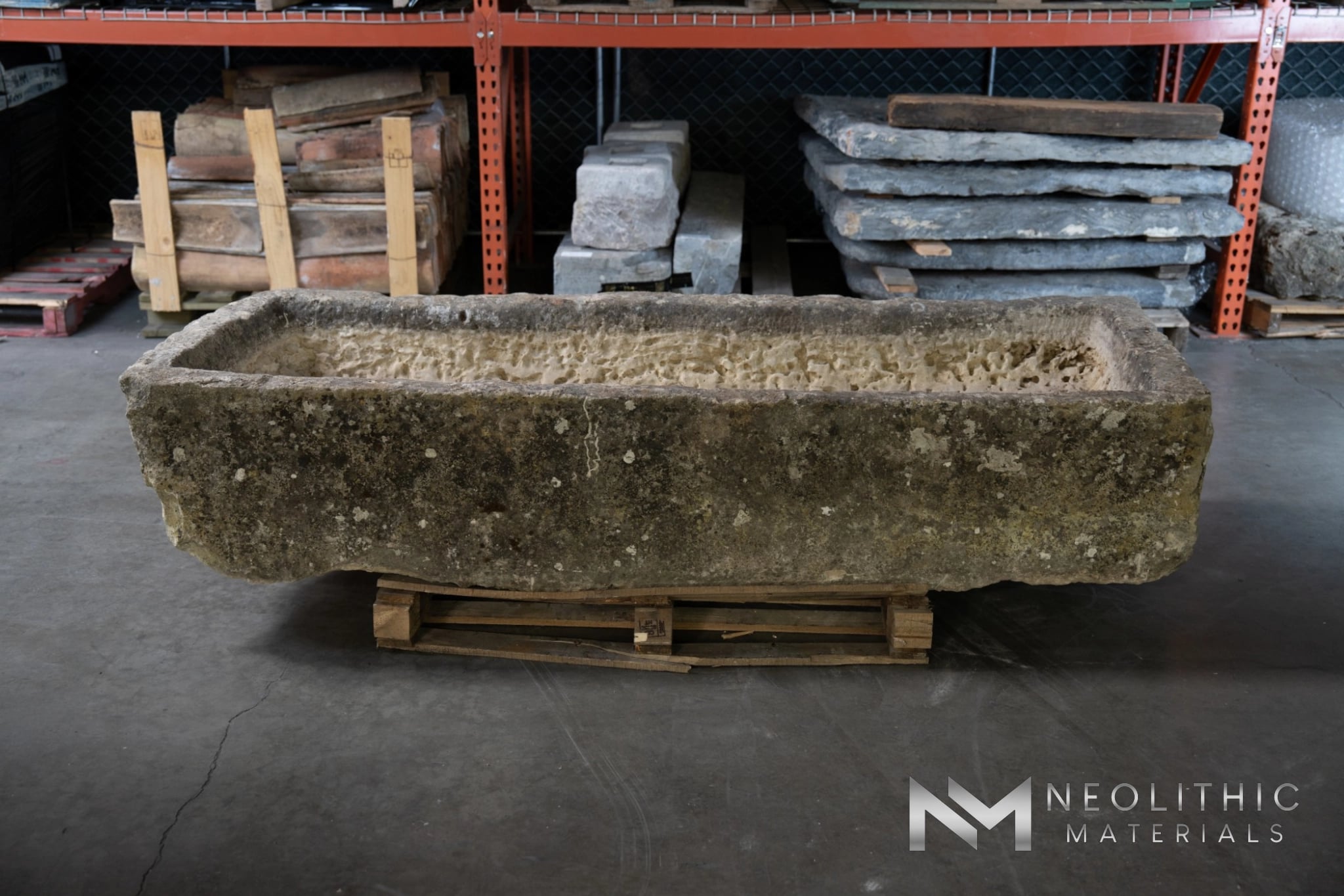 Trough FG103-21.MN.87x33x17 wide warehouse storage shot showcasing a massive, rectangular antique stone trough with a heavily weathered, mossy dark brown patina, elevated on a wooden shipping pallet with stone slabs stacked in the background.