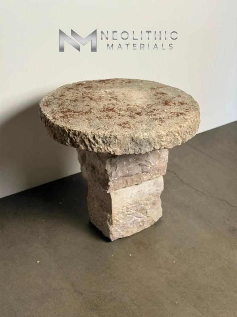Tables and Slabs FG104-01.28x3 eye-level indoor showroom photograph showcasing a rustic round stone table with a heavily textured, pitted limestone top supported by a thick, square chiseled pedestal base on a concrete floor.
