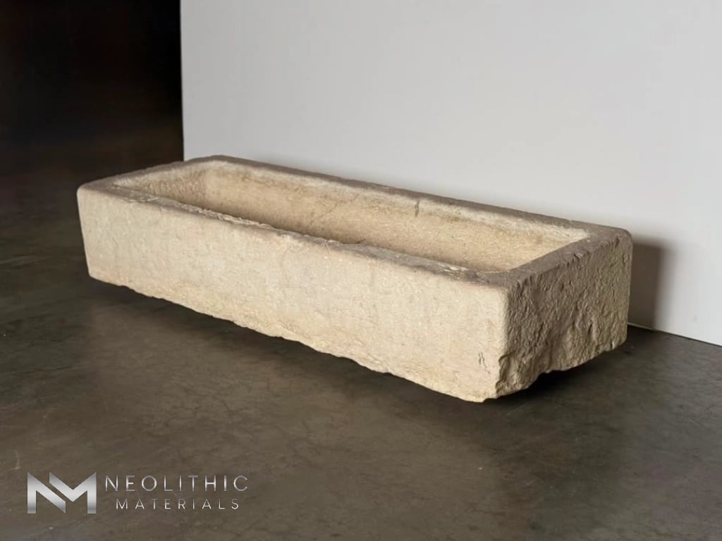 Trough FG104-23.56x20x11 eye-level studio photograph showcasing a long, rectangular chiseled stone block trough featuring a deeply pitted beige limestone exterior texture, positioned on a dark polished concrete surface against a white wall.