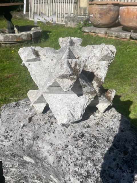 Miscellaneous FI103-05.LY Reclaimed multi-faceted geometric stone finial fragment featuring intricate chiseled triangular planes and a light grey weathered finish, captured in a bright outdoor close-up shot balanced on a large textured boulder.