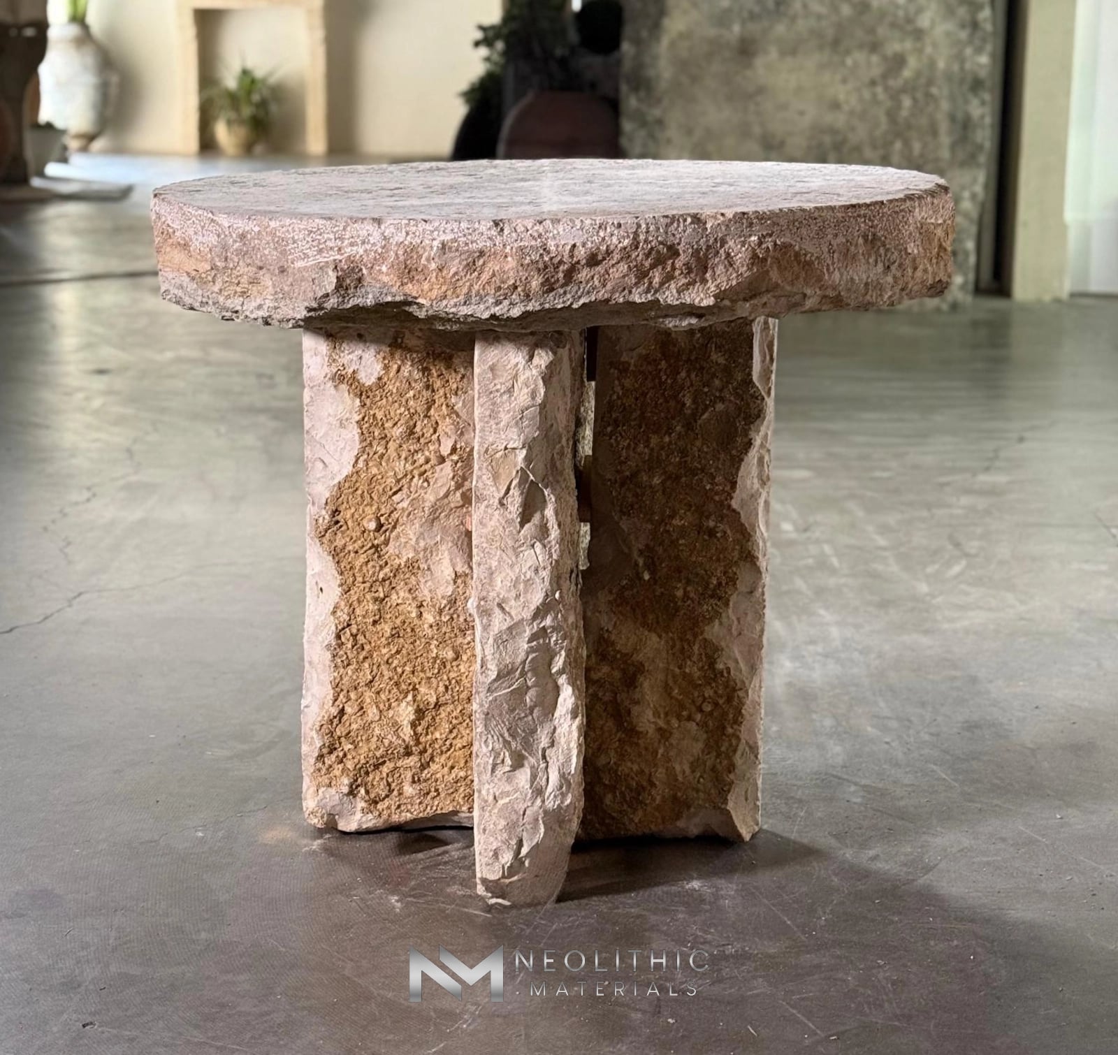 Tables and Slabs FK103-72: Medium shot of a rustic round table crafted from heavily textured, natural beige stone, featuring a thick hand-chiseled top supported by an intersecting slab pedestal base inside a showroom setting.