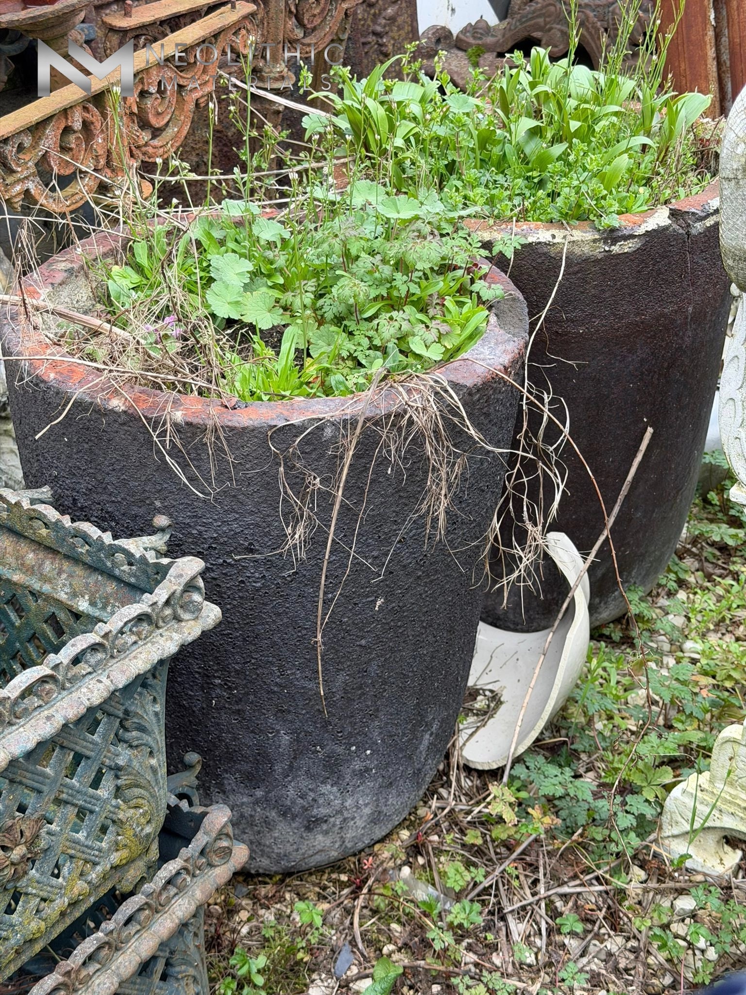 Pots and Planters FK104-06 & FK104-07 ground-level exterior view highlighting a pair of matching tall, tapered antique stone vessels with heavily distressed charcoal-colored exterior finishes and contrasting smooth terra-cotta toned rims nestled among overgrown grass and garden salvage elements.