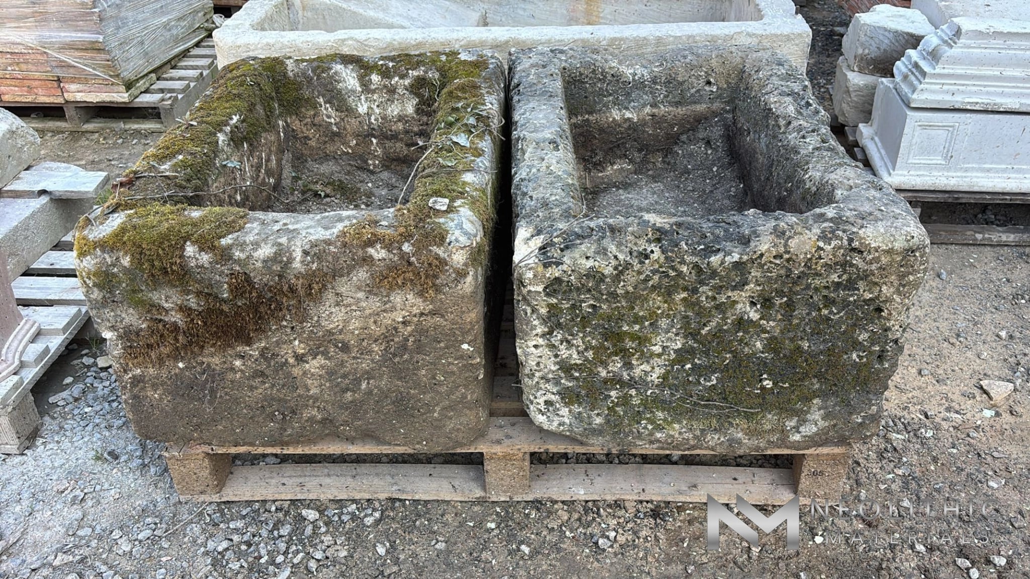 Trough FS101-03.M & FS101-04.M straight-on eye-level view focusing on two thick, square-shaped antique stone planters sitting adjacent on a shipping pallet, emphasizing their green moss growth and heavily distressed, ancient chiseled surfaces.