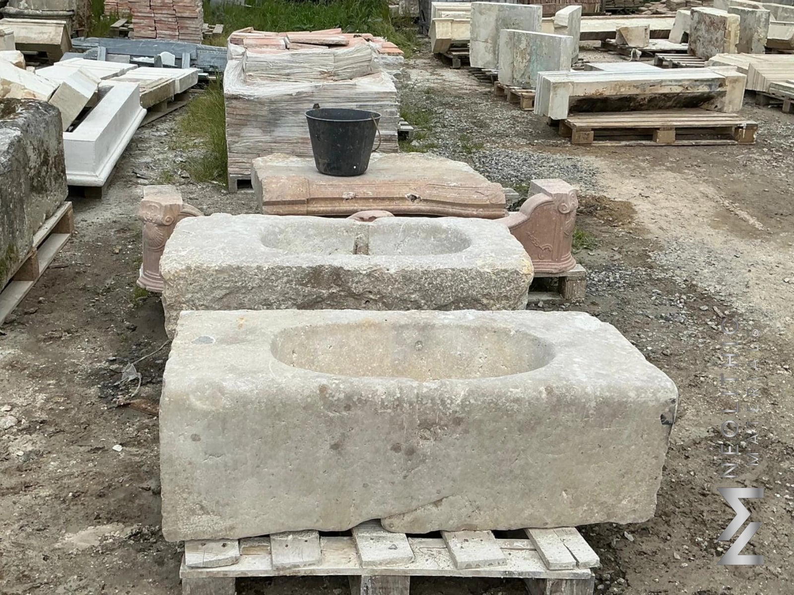 Trough FS101-07.MN & FS101-08.MN wide-angle outdoor perspective shot showcasing a row of heavy rectangular blocks of carved limestone with smooth circular basins hollowed out of their tops, resting on pallets in a gravel stone supply yard.