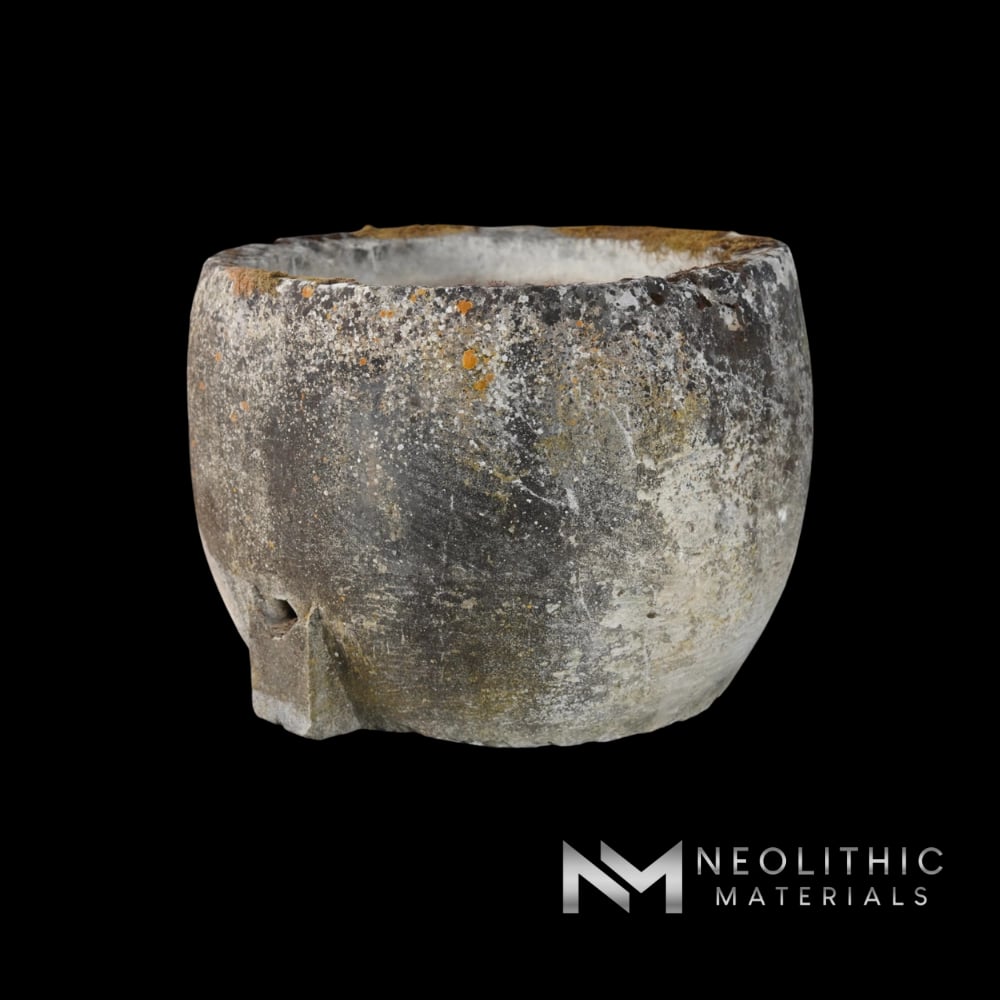 Trough FS101-09.Y wide outdoor shot highlighting a massive, cup-shaped antique stone basin with a dark, deeply weathered surface and a singular raised drainage spout at the lower rim, resting on a wooden storage pallet.