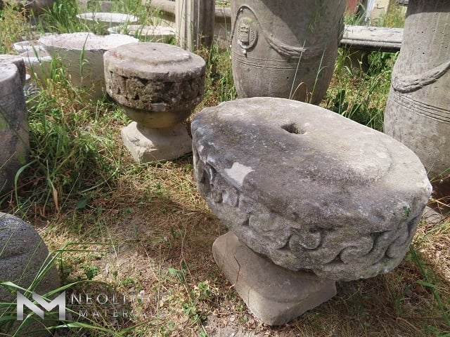 Miscellaneous FU101-01 & FU101-02: Side-angle courtyard view displaying two round, antique carved stone architectural blocks with highly textured relief flourishes, repurposed as rustic garden pedestal elements resting on natural soil and sparse grass.