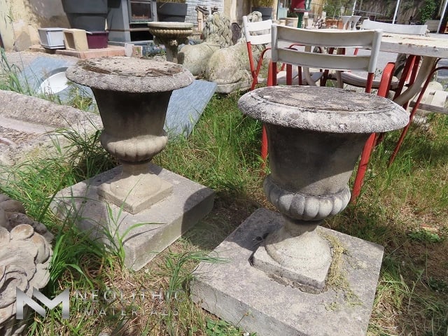 Miscellaneous FU101-03 & FU101-04: Ground-level outdoor picture displaying two antique cast stone garden planters with fluted goblet shapes and heavily patinated textures, set sequentially on concrete display blocks against a background of rustic terrace patio furniture.