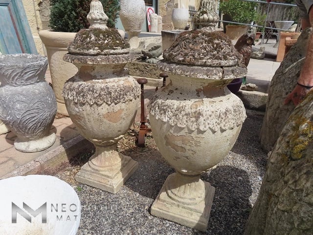 Miscellaneous FU101-05 & FU101-06: High-angle exterior photograph showcasing a pair of massive, classical stone garden urns with matching finial lids, featuring highly weathered surfaces covered in dark moss and intricate leaf relief carvings on a gravel patio floor.