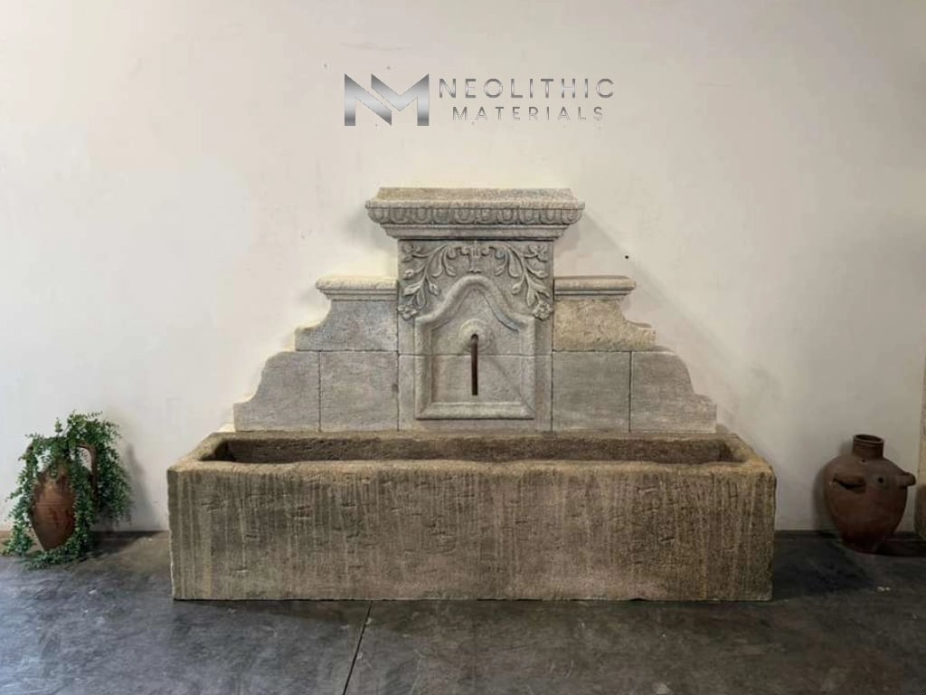 Fountain LA343-01 & LA343-02: Centered indoor snapshot of a grand antique stone wall fountain featuring an ornate stepped stone backing with carved scrollwork, centered above a long weathered rectangular trough basin resting on a concrete showroom floor.