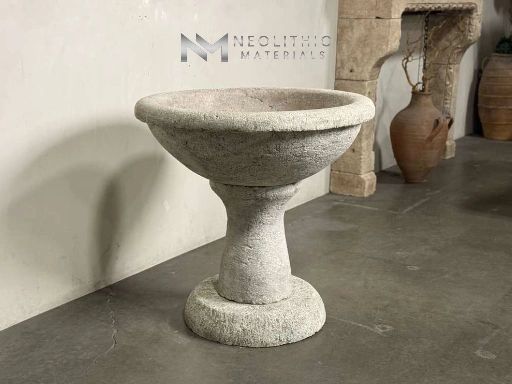 Fountains LA343-05.36x4x36 eye-level showroom display of a classic stone pedestal basin fountain, featuring a deeply pitted, weathered white concrete texture and an elegant chalice profile resting on a dark concrete floor.